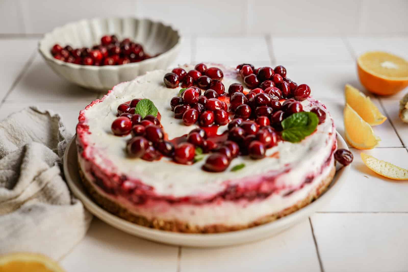 No-bake vegan cheesecake with cranberries on top, surrounded by oranges and a bowl of fresh cranberries on countertop.