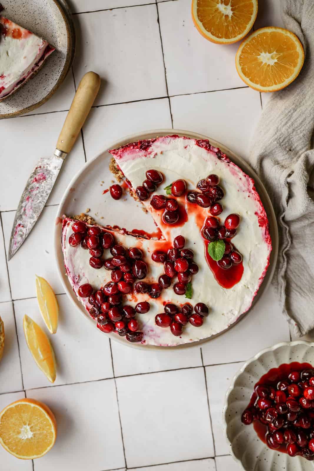 No-bake vegan cheesecake with cranberries and raspberries on top, surrounded by oranges on counter, with slice out of it.