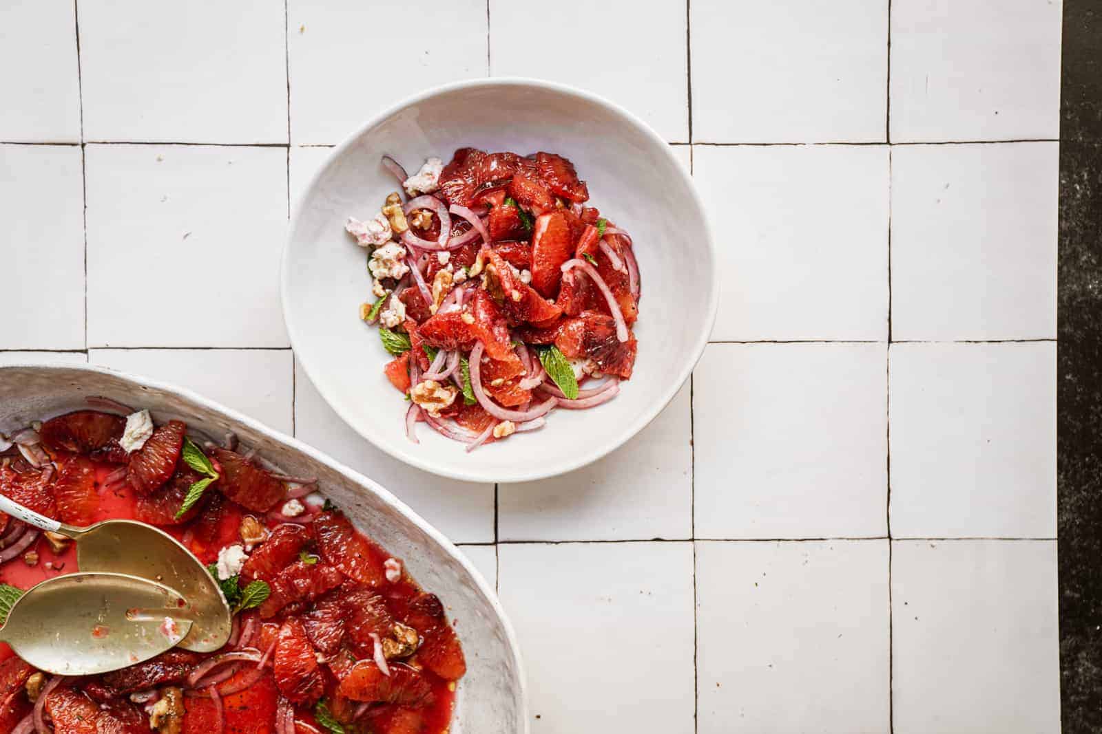 Colorful blood orange salad in a bowl next to a large salad bowl on white countertop