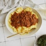 A bowl of wide ribbon pasta topped with a rich, chunky vegan ragu. A gold fork rests on the bowls edge. A white cloth napkin, a sprig of herbs, and a bottle of olive oil are nearby on a white tiled surface.