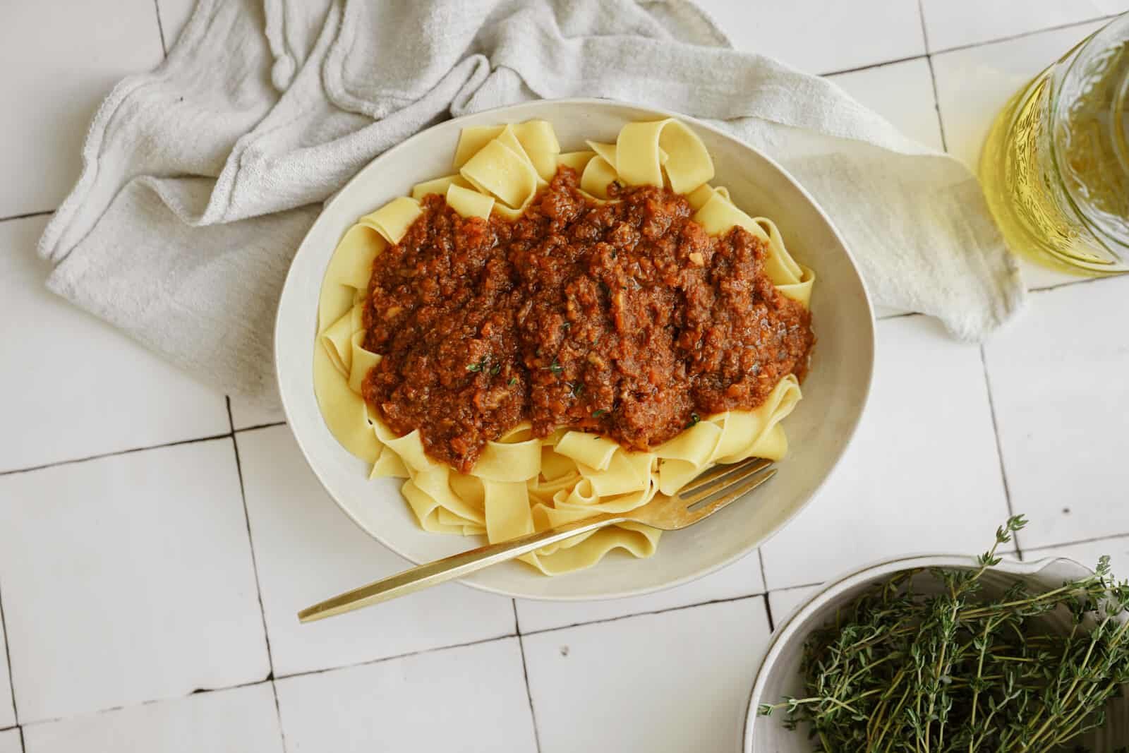 A bowl of wide ribbon pasta topped with a rich, chunky vegan ragu. A gold fork rests on the bowls edge. A white cloth napkin, a sprig of herbs, and a bottle of olive oil are nearby on a white tiled surface.