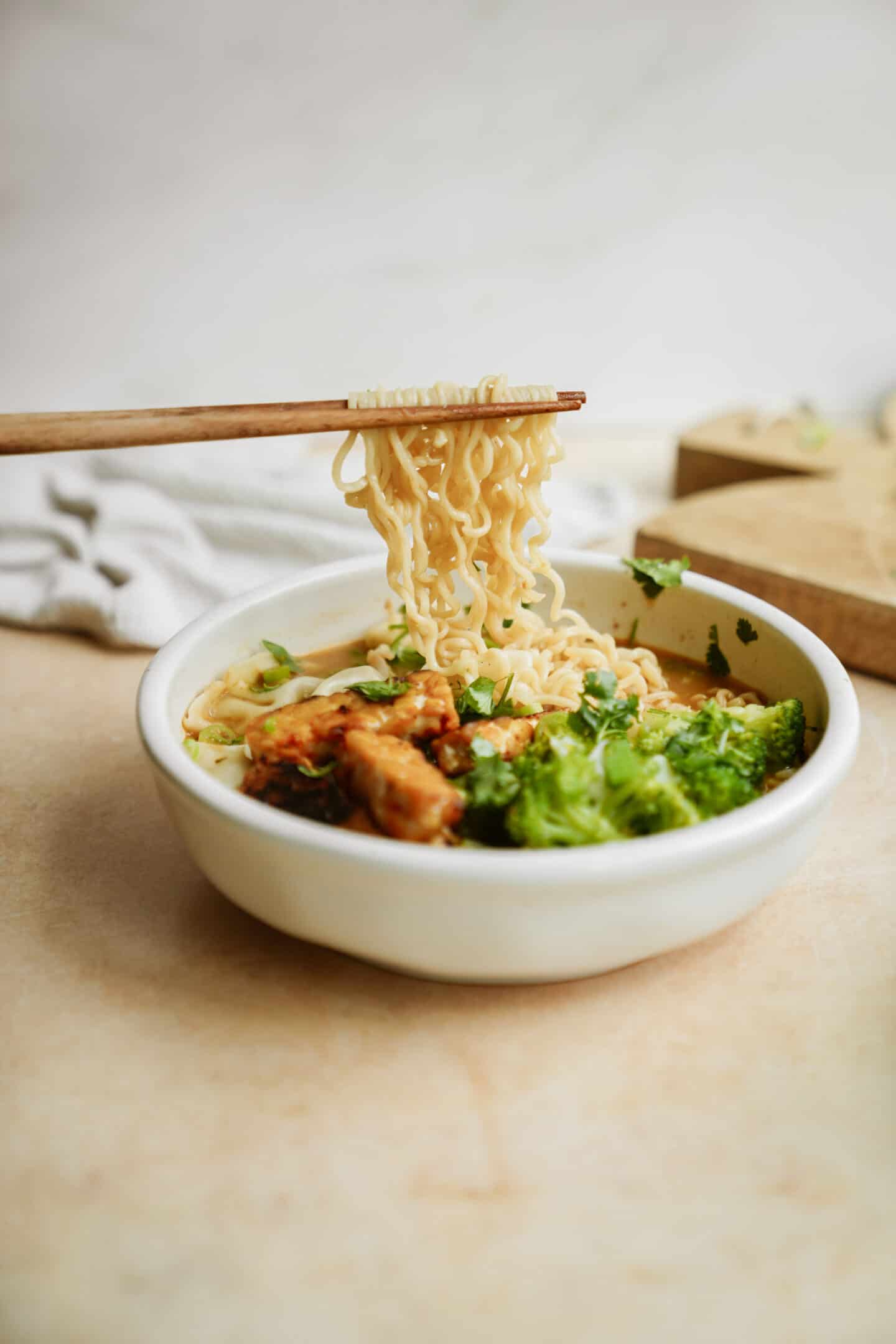 A white bowl filled with creamy vegan ramen, showcasing noodles topped with pieces of meat and broccoli. A pair of chopsticks gracefully lifts a portion, while a light-colored napkin and wooden board set the background scene.