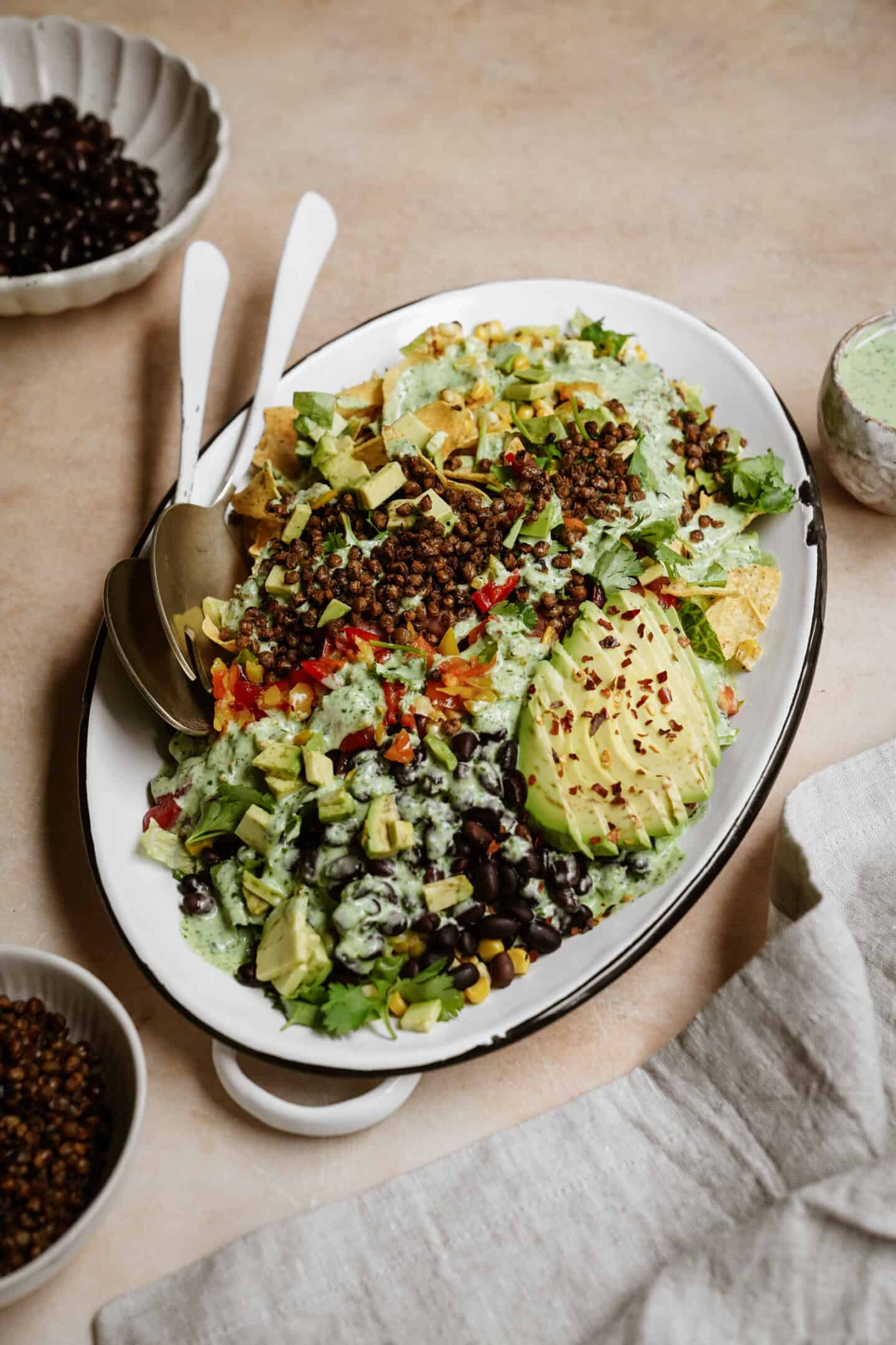 A vibrant platter of vegan nachos transformed into a vegan taco salad, topped with lentils, black beans, avocado slices, chopped lettuce, and diced red peppers, all drizzled with green sauce. Nearby are bowls of lentils and creamy sauce with a fabric napkin beside the plate.