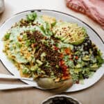 A white oval platter brimming with a Vegan Taco Salad features nachos topped with lentils, sliced avocado, black beans, corn, diced tomatoes, and greens drizzled in green sauce. A cloth napkin sits beside the platter, while a serving spoon and fork rest invitingly on the dish.
