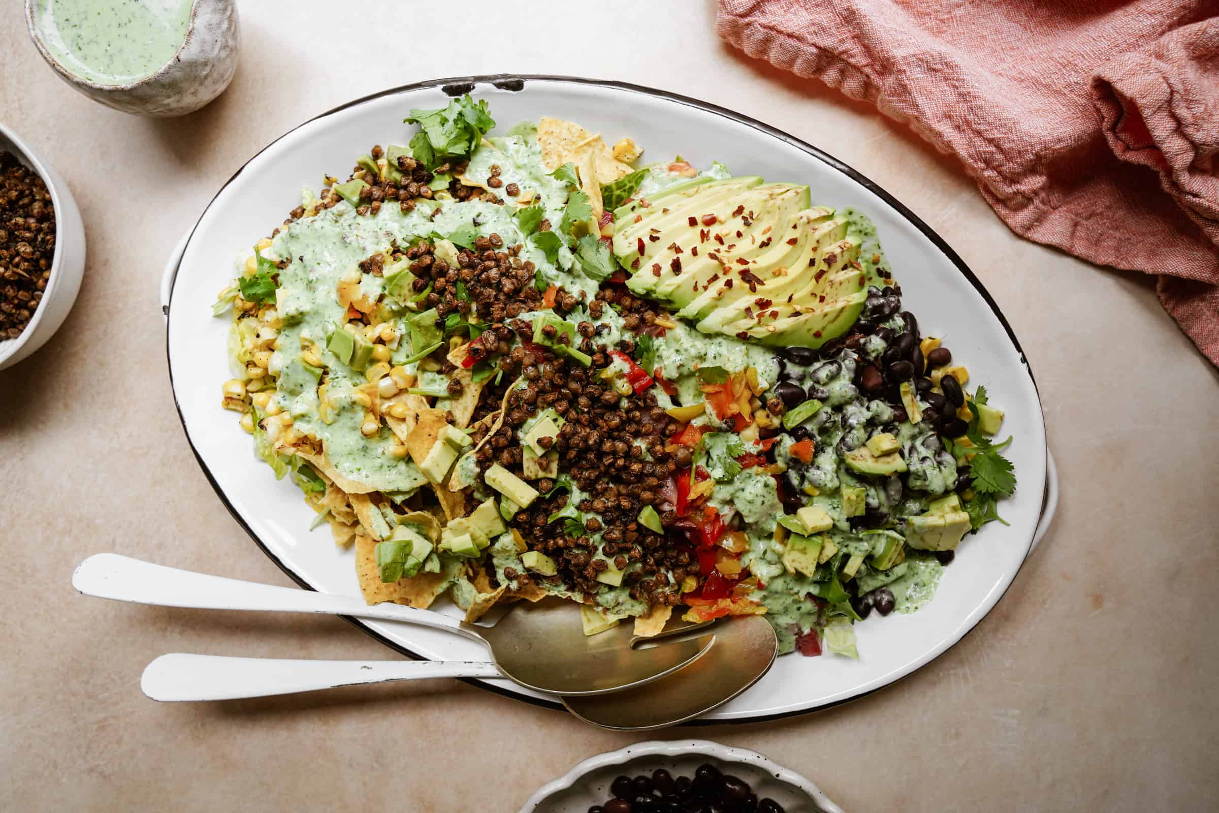 A white oval platter brimming with a Vegan Taco Salad features nachos topped with lentils, sliced avocado, black beans, corn, diced tomatoes, and greens drizzled in green sauce. A cloth napkin sits beside the platter, while a serving spoon and fork rest invitingly on the dish.
