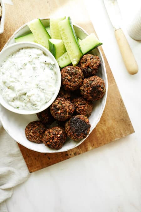A bowl featuring falafel, cucumber sticks, and a small bowl of creamy tzatziki sauce sits on a wooden board with a knife nearby—perfect inspiration for Walnut Meatballs with Tzatziki Sauce.