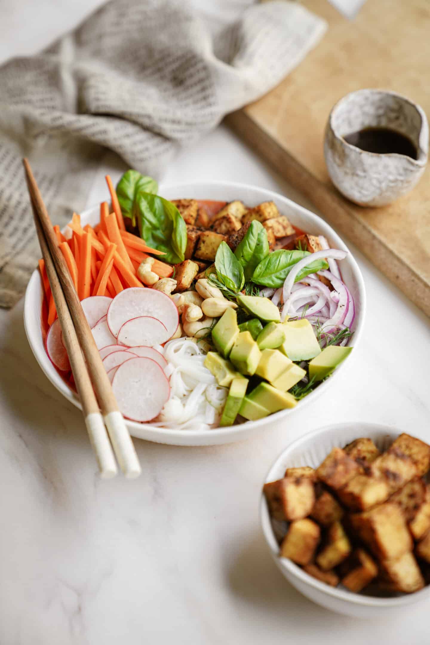 A colorful bowl of sliced vegetables, tofu, avocado, and herbs with chopsticks on the side. Nearby is a small bowl of cubed tofu and a cup of sauce. A napkin and wooden board are in the background.