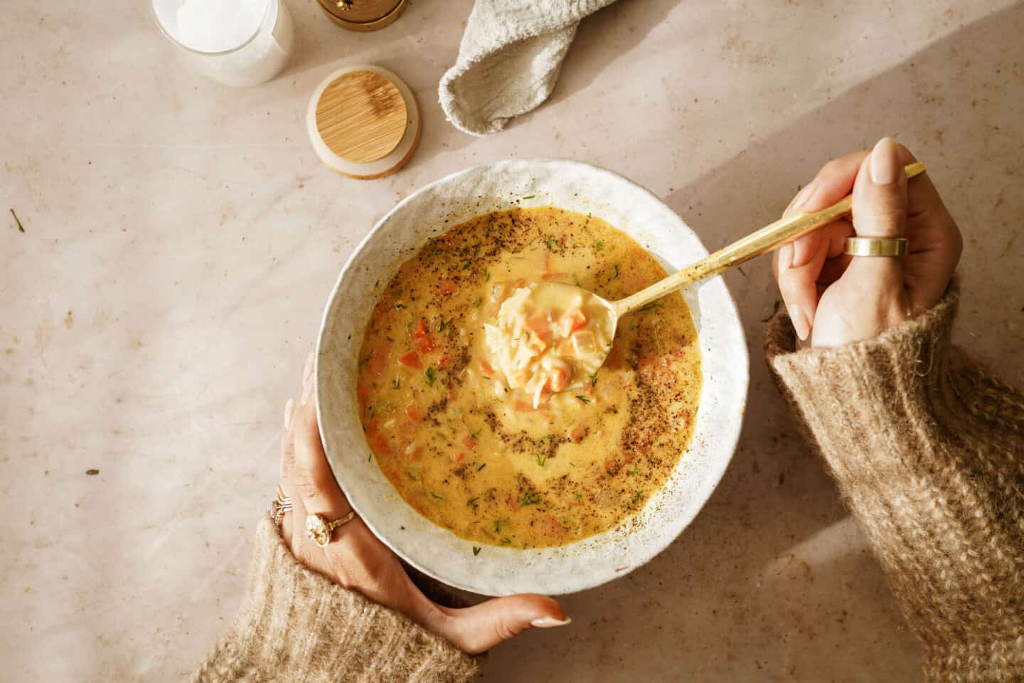 A person in a cozy sweater holds a bowl of vegan Avgolemono soup, garnished with fresh herbs. They are about to savor a spoonful. The scene is warmly lit, with a soft, rustic background.