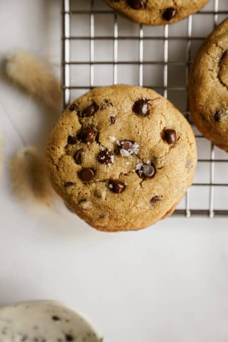 A chocolate chip cookie sprinkled with flaky sea salt rests on a metal cooling rack, with part of another cookie visible nearby and a blurred white background.
