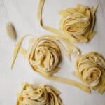 Four nests of uncooked, homemade fettuccine pasta rest on a white surface, with some loose strands scattered around them. A metal rack and a dried decorative plant are partially visible at the top—perfect for your next vegan pasta recipe.