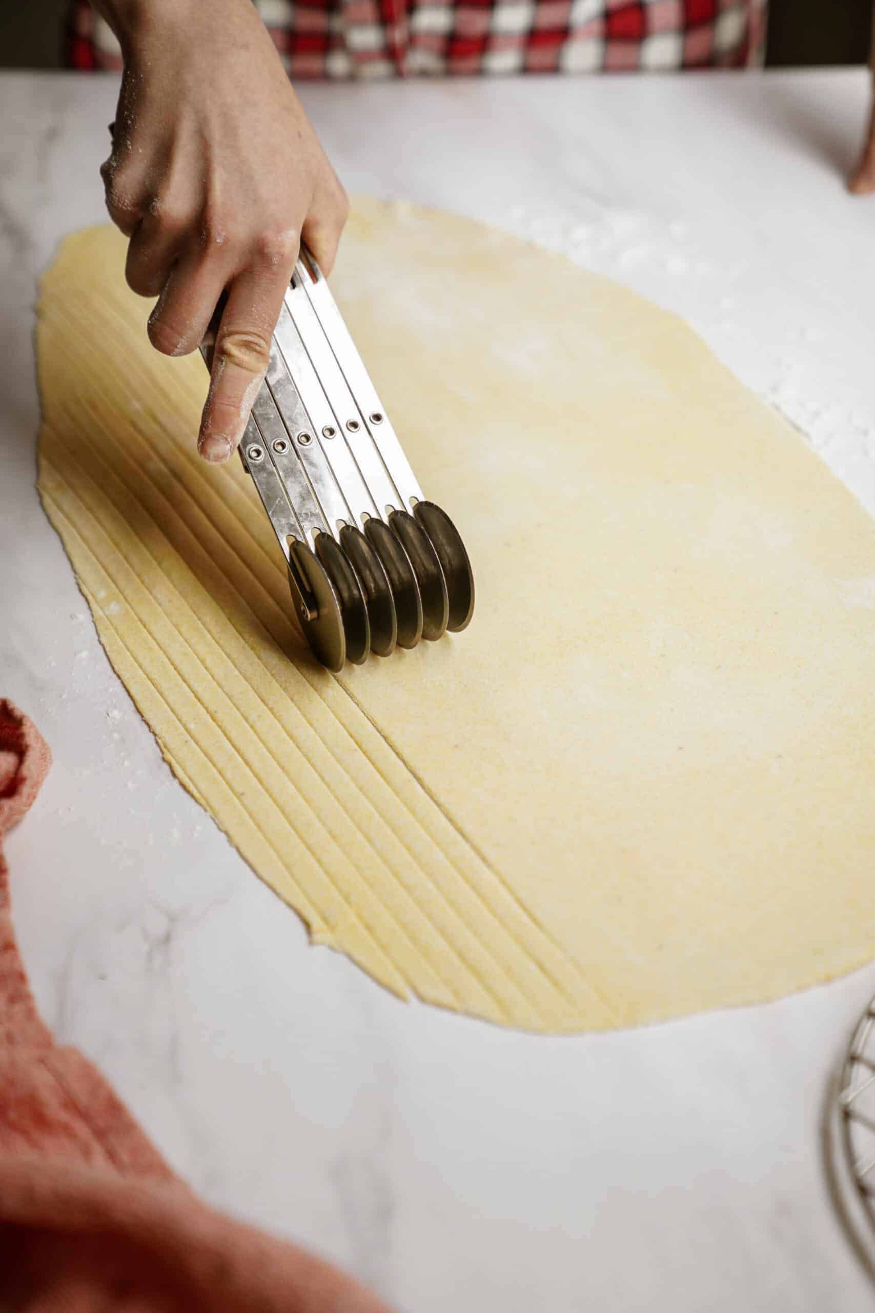 A hand uses a multi-wheel dough cutter to slice thin, even strips from a large sheet of rolled-out dough on a floured surface, perfect for creating homemade vegan pasta recipes.