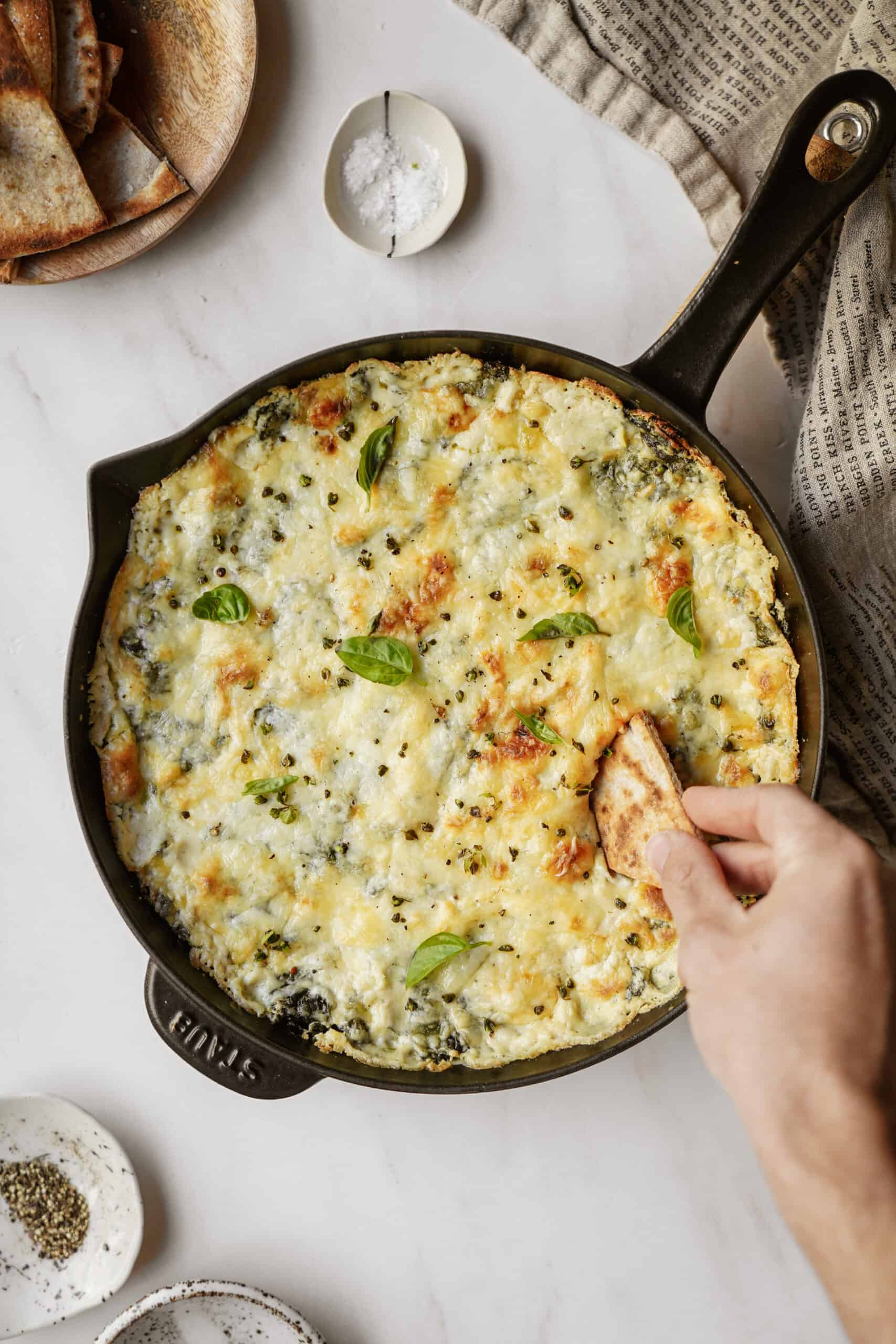 Spinach and Artichoke Dip in cast iron dish with hand dipping pita chip in