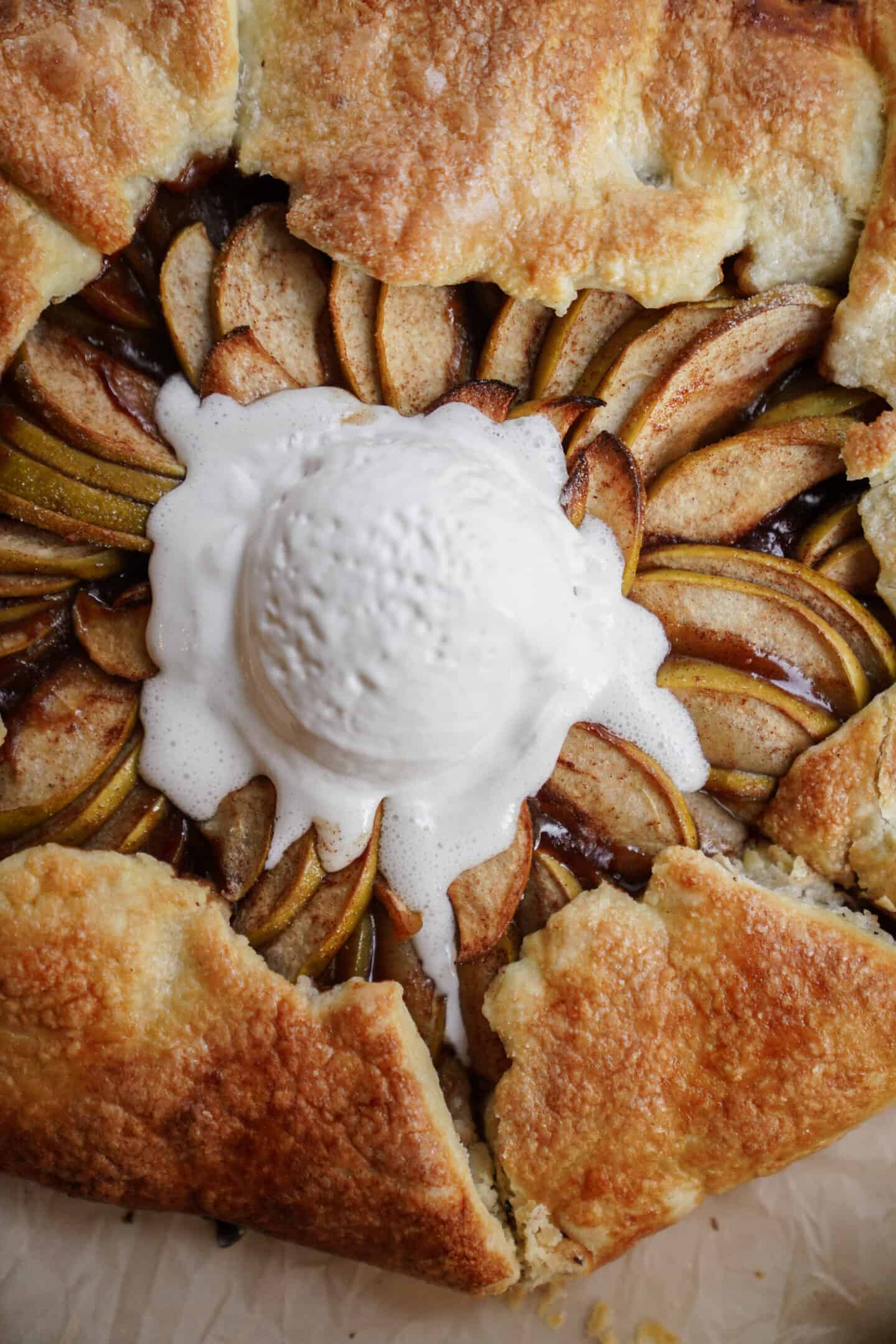 A close-up of an Apple Galette with golden, flaky crust, thinly sliced apples arranged in a circular pattern, and a scoop of melting vanilla ice cream in the center.
