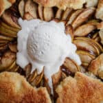 A close-up of an Apple Galette with golden, flaky crust, thinly sliced apples arranged in a circular pattern, and a scoop of melting vanilla ice cream in the center.