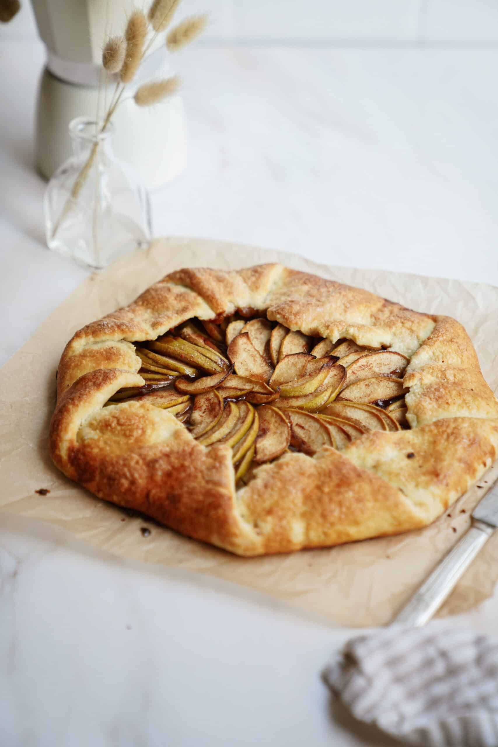 A rustic Apple Galette with a golden, flaky crust and neatly arranged apple slices sits on parchment paper. A knife and striped cloth rest nearby, while a glass vase with dried grasses adds charm in the background.