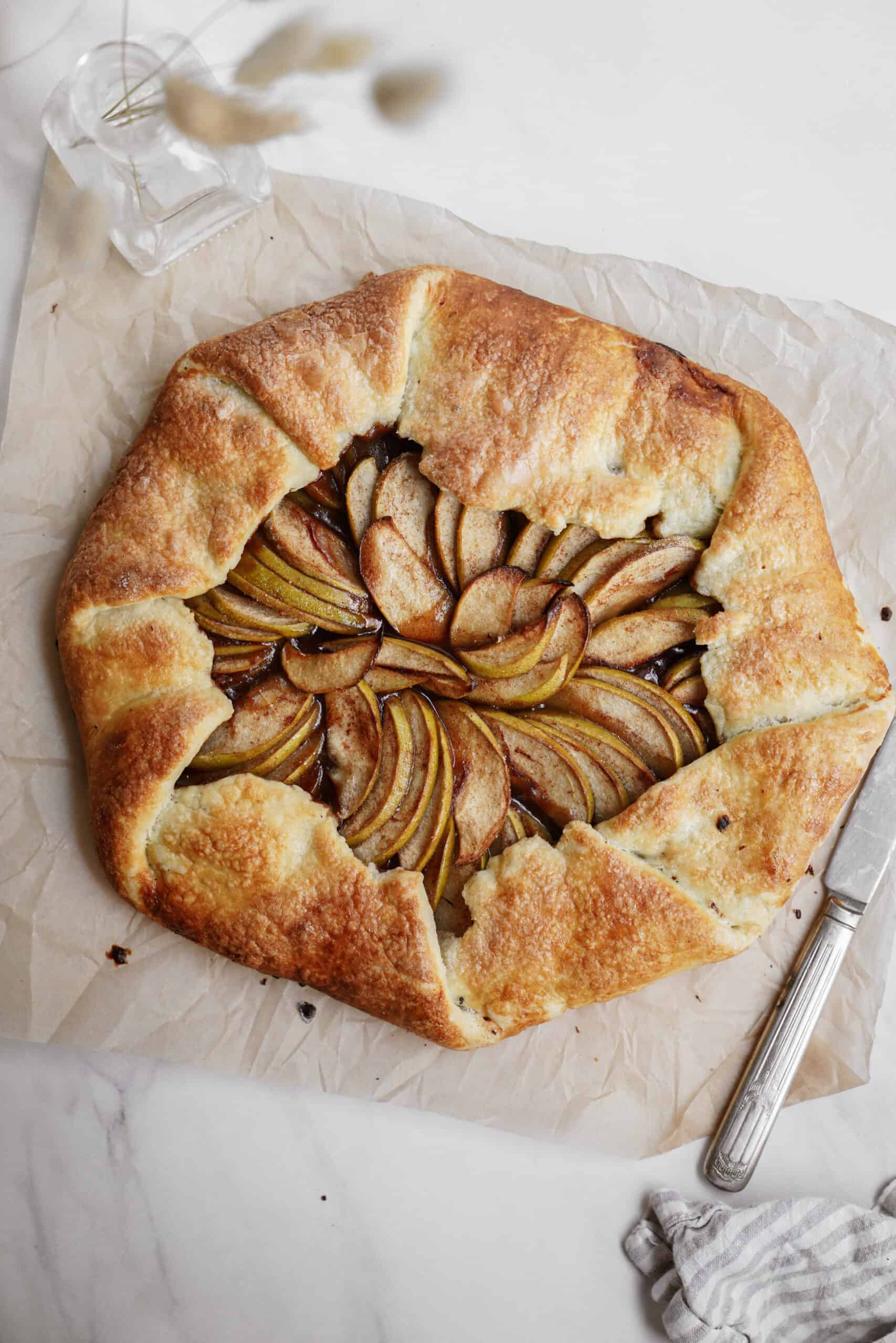 A golden-brown apple galette with a rustic, folded crust sits on parchment paper. The sliced pears and apples are arranged in a spiral pattern. A knife rests nearby on the marble surface.