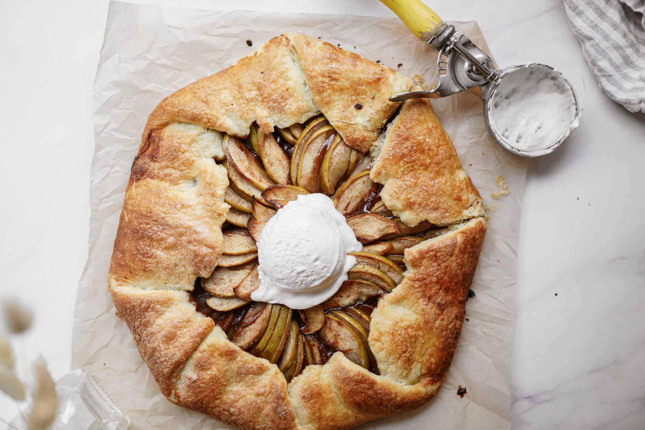 A rustic Apple Galette with a golden, flaky crust is topped with a scoop of vanilla ice cream. An ice cream scoop with remnants of the sweet treat rests nearby on a sheet of parchment paper.