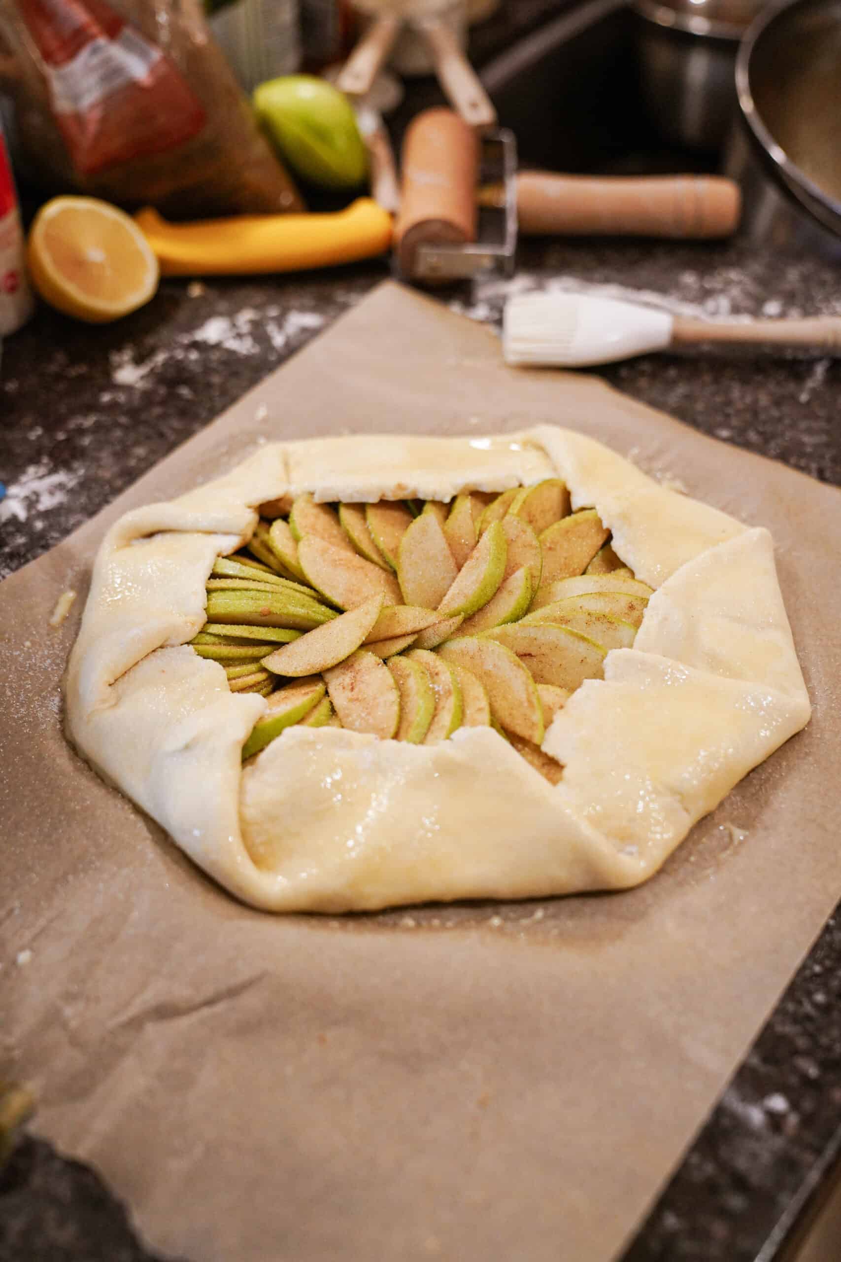 An unbaked rustic Apple Galette with folded edges sits on parchment paper, ready for the oven. Sliced apples are neatly arranged inside the pastry, surrounded by baking tools and ingredients on a dark countertop.
