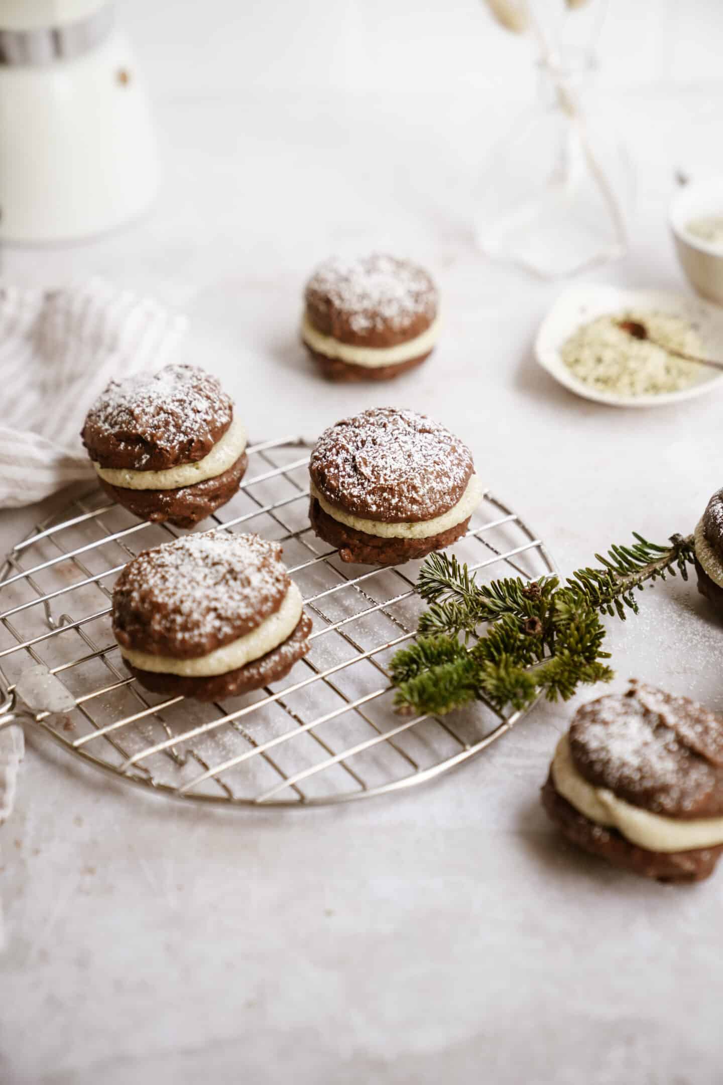 Five chocolate whoopie pies with cream filling, made from a classic whoopie pie recipe, are arranged on a cooling rack and dusted with powdered sugar. A pine sprig rests nearby, with baking ingredients in the background.