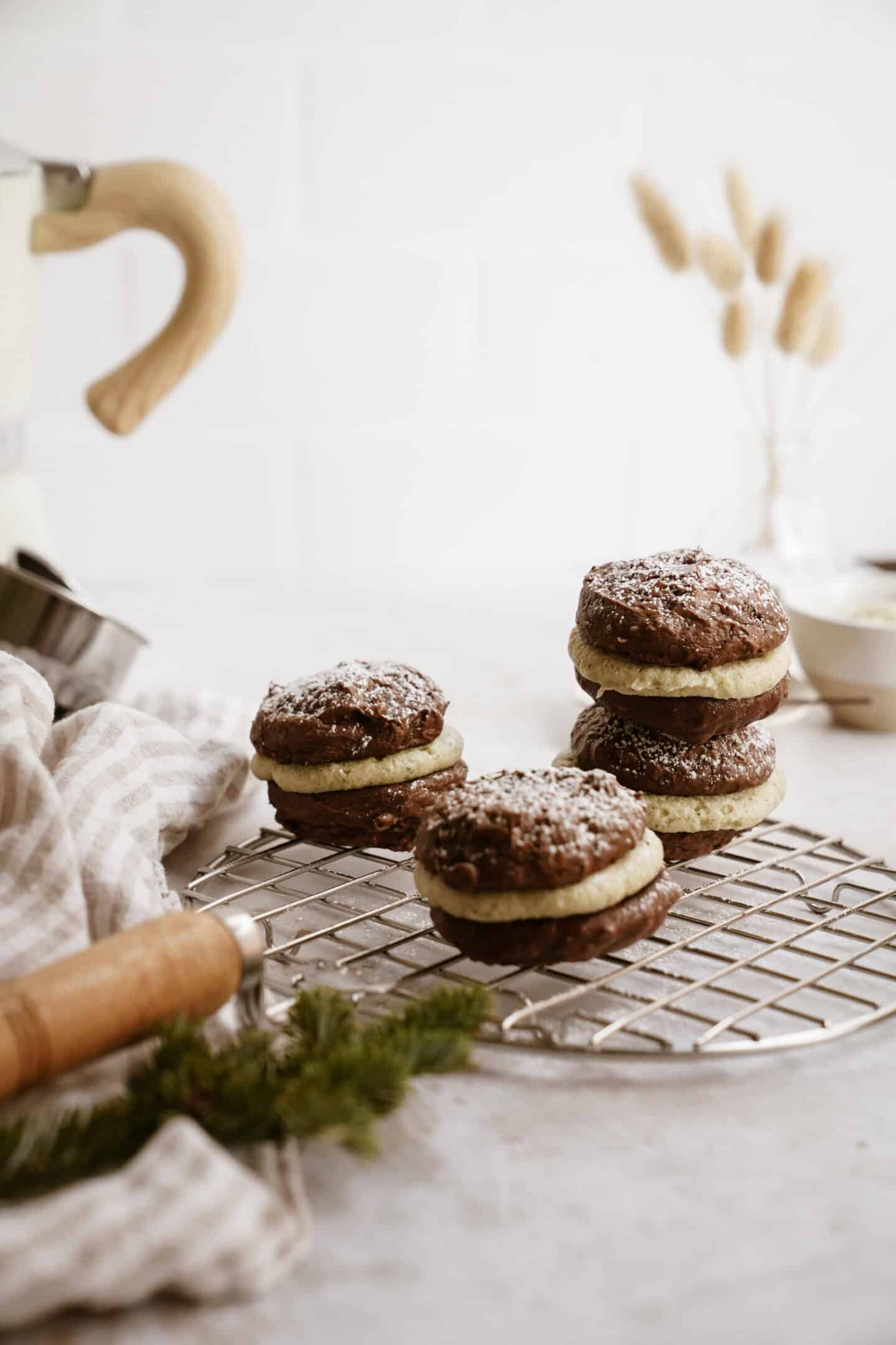 Four chocolate whoopie pies with cream filling, dusted with powdered sugar, are stacked on a round cooling rack. A striped cloth and kitchen utensils sit nearby—a tempting showcase for your next whoopie pie recipe.