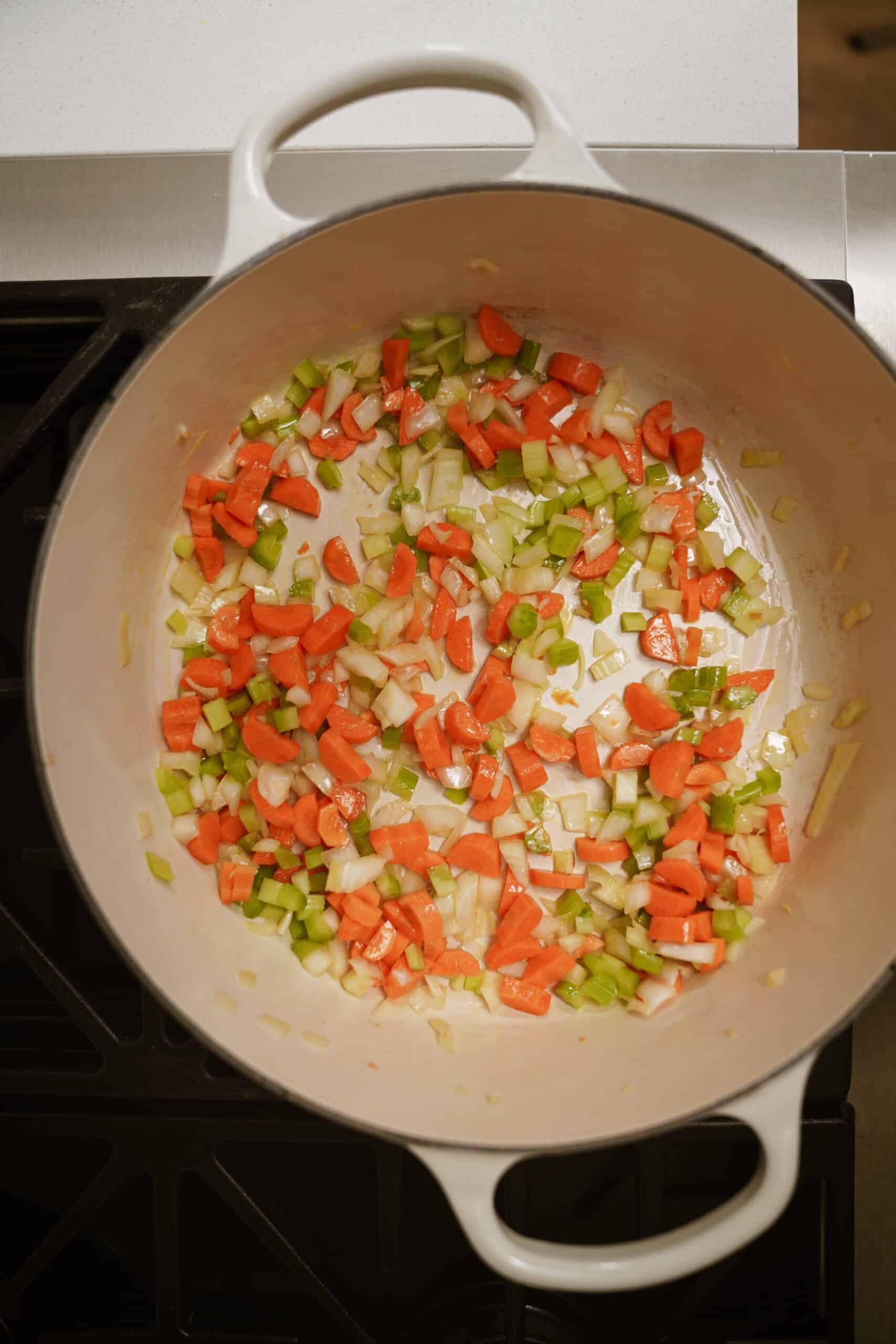 Chopped carrots, celery, and onions are being sautéed in a large white pot on a stove, forming the flavorful base for a comforting cheeseburger soup.