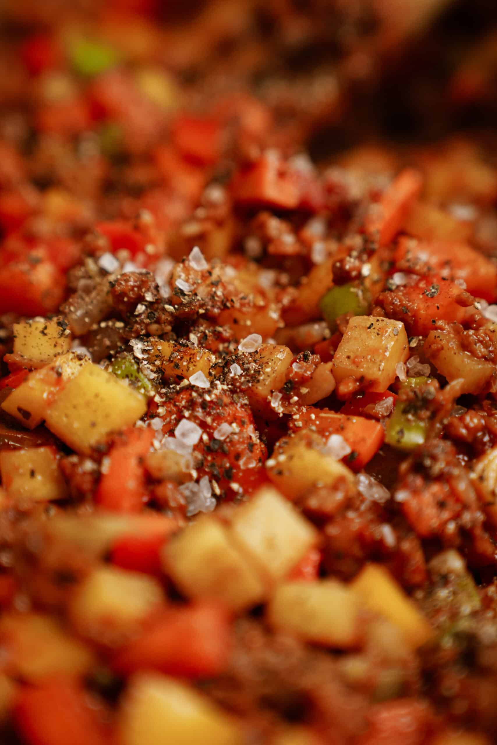 Close-up of a colorful mixture of diced vegetables and ground meat, seasoned with black pepper and coarse salt. This vibrant blend, reminiscent of hearty cheeseburger soup, features freshly cooked red, green, and yellow ingredients.