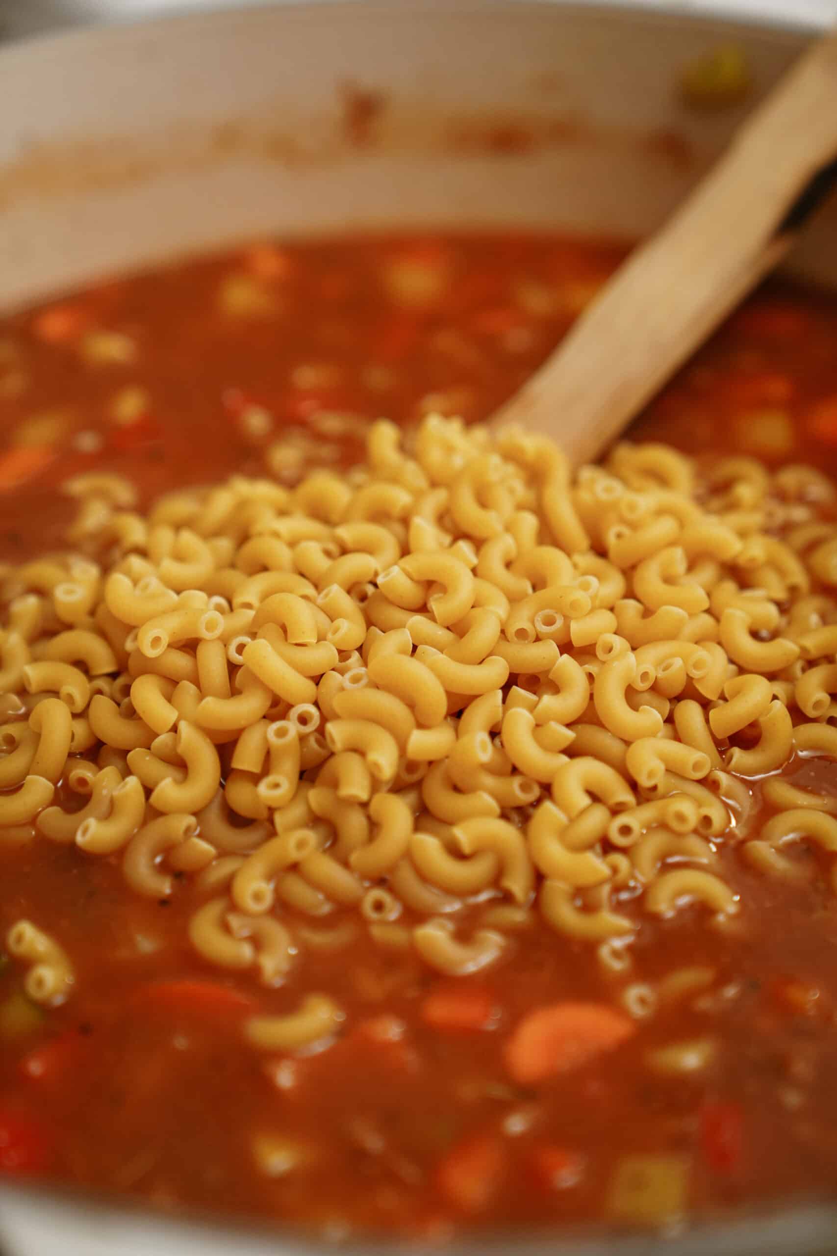 Uncooked elbow macaroni being stirred into a pot of cheeseburger soup with a wooden spoon; the reddish broth features visible chopped vegetables.