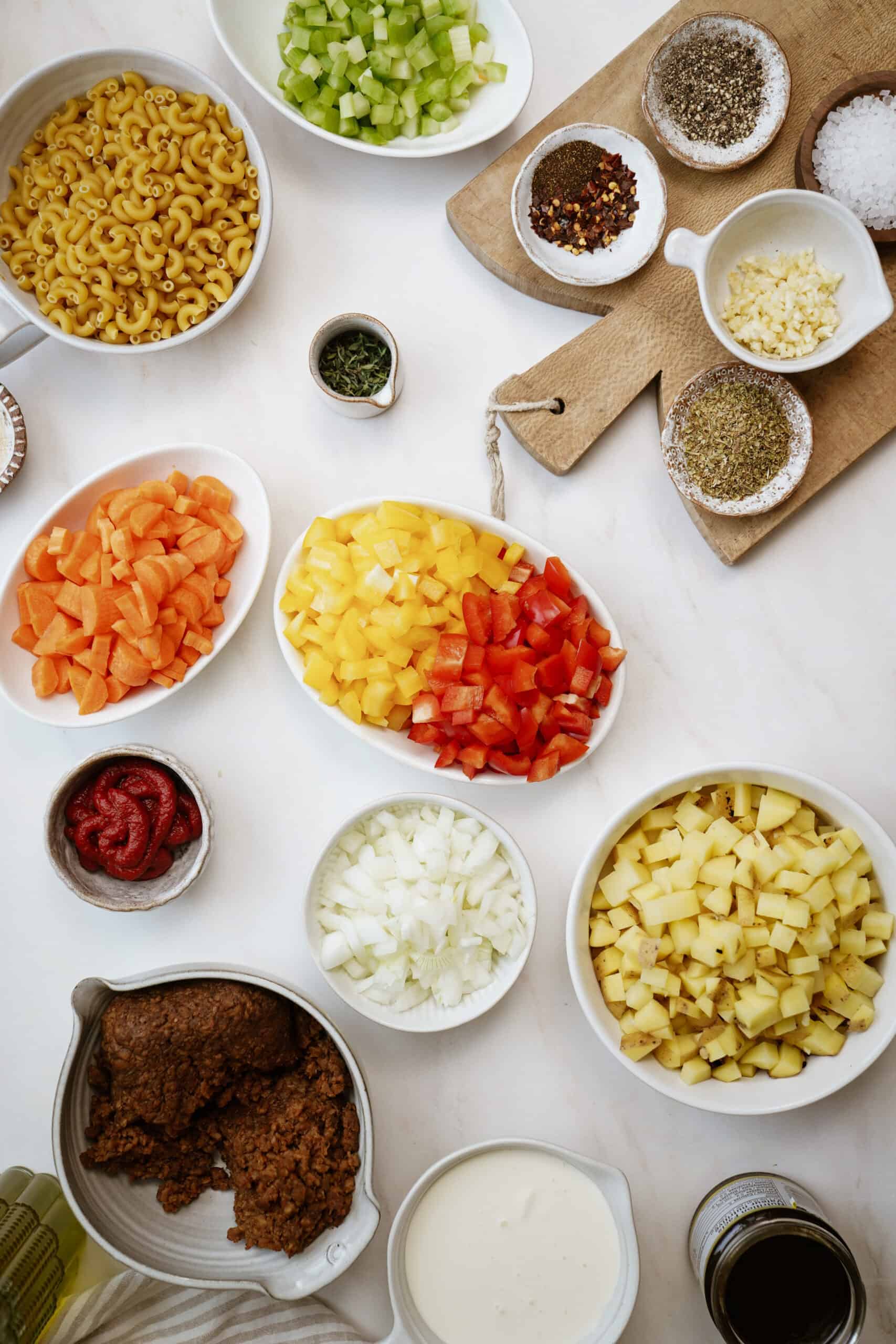 Various bowls and dishes containing chopped vegetables, macaroni, tomato paste, spices, plant-based meat, and cream are arranged neatly on a white surface, ready for cooking a hearty batch of plant-based cheeseburger soup.