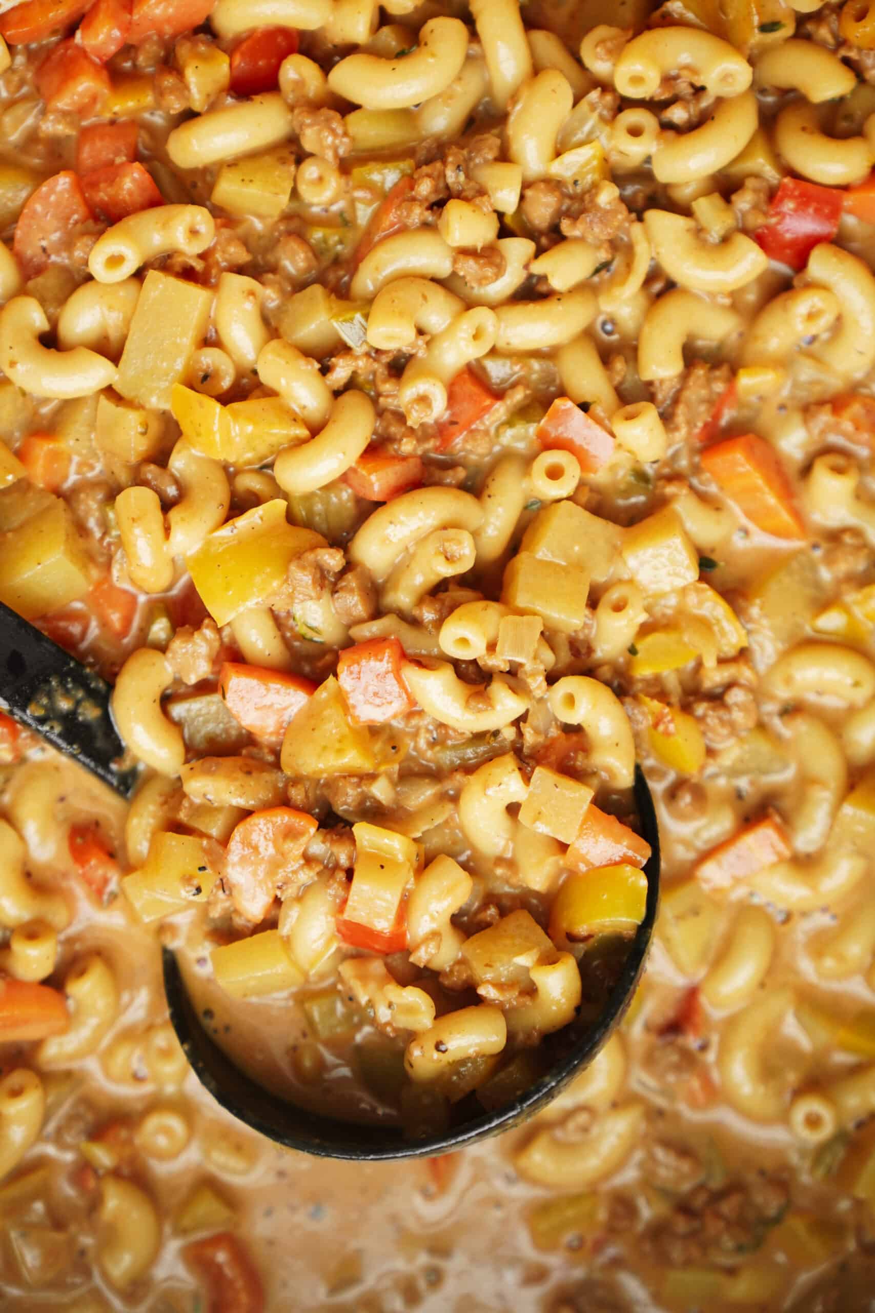 Close-up of creamy cheeseburger soup with macaroni, ground meat, diced carrots, potatoes, and celery, shown in a ladle and spread throughout the pot.