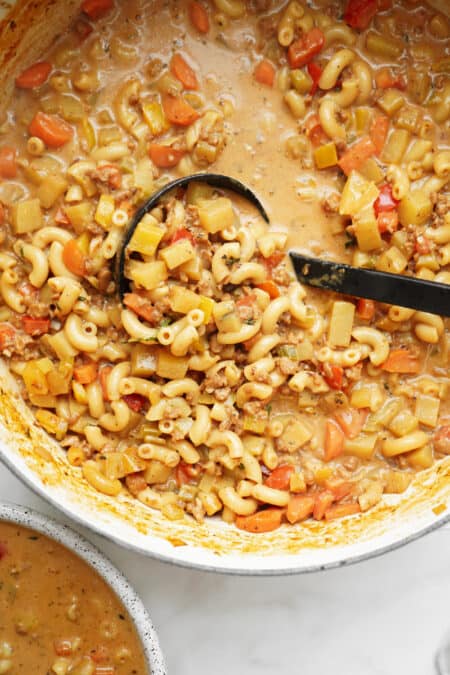 A ladle scoops up hearty macaroni cheeseburger soup with diced vegetables, ground meat, and a creamy broth from a large pot. The soup features elbow pasta, carrots, celery, and tomatoes.