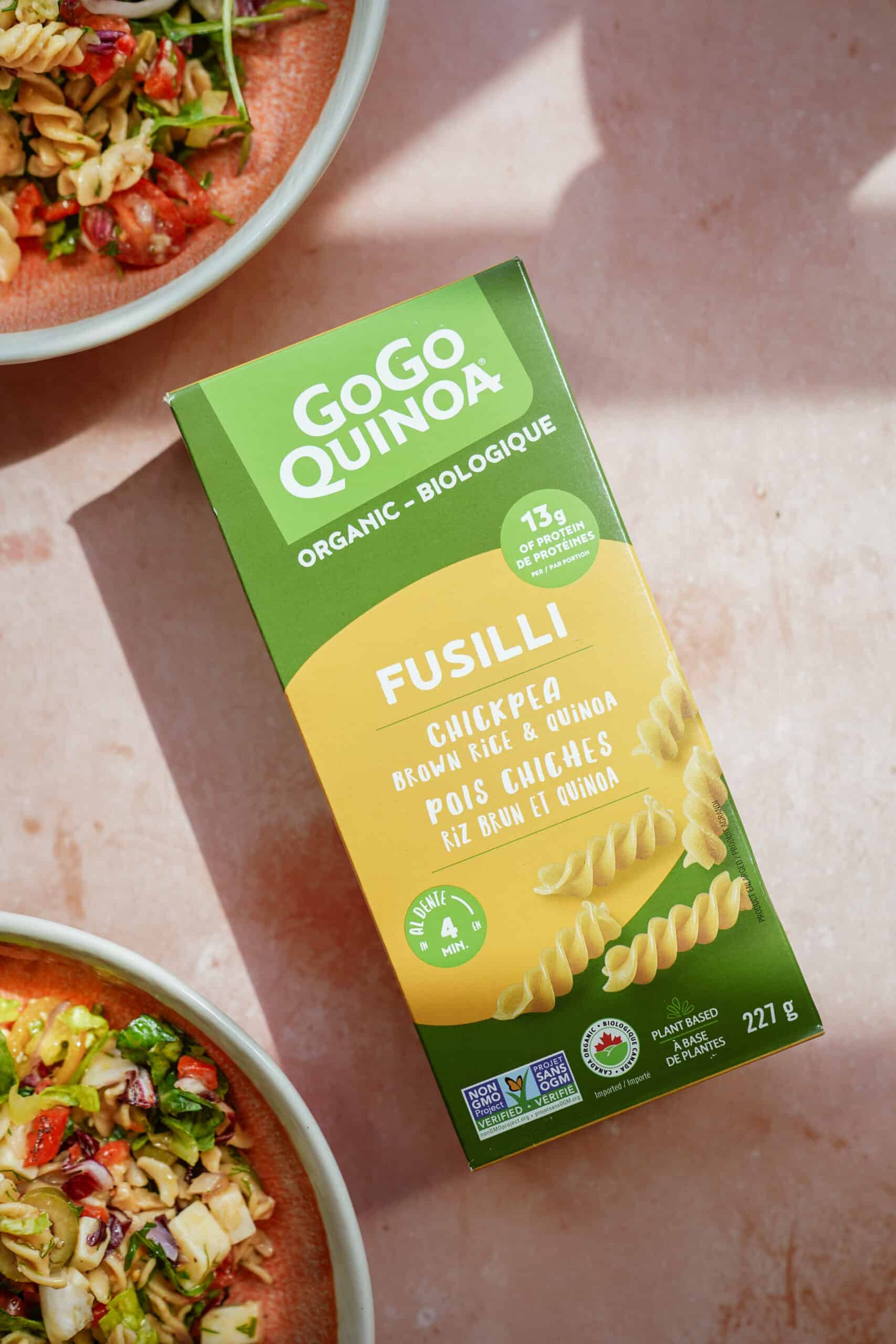 A box of GoGo Quinoa organic fusilli chickpea pasta sits on a pink surface, perfect for making a nutritious Chickpea Pasta Salad. Partially visible salad bowls with fresh vegetables appear in the background.
