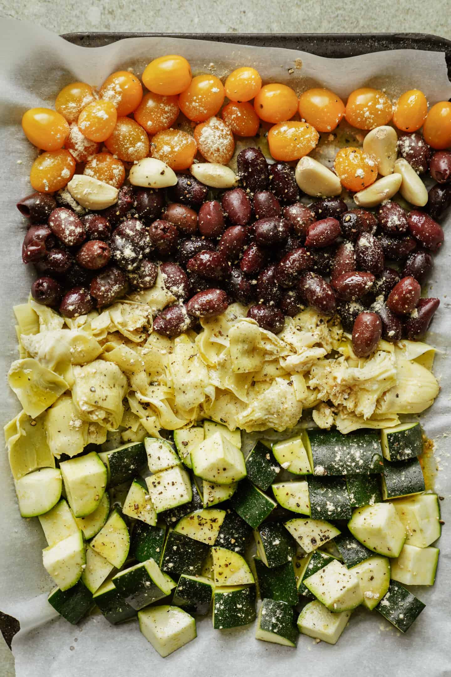 A baking sheet lined with parchment paper holds rows of yellow cherry tomatoes, garlic cloves, Kalamata olives, marinated artichoke hearts, and cubed zucchini—like a rustic pasta primavera—sprinkled with seasoning and ready to be roasted.