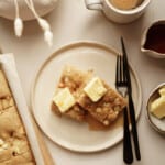 A plate with two pieces of Sheet Pan Pancakes (High Protein), topped with butter and syrup, sits beside a mug of coffee, a small jug of syrup, a bowl of butter, and a baking tray on a light table. Black fork and knife rest on the plate.