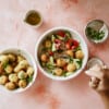 A hand holds a fork over a bowl of salad with cherry tomatoes, cucumbers, onions, and crispy bread pieces. Nearby are a bowl of Feta Stuffed Olives (fried), a small bowl of herbs, and a jug of olive oil on a pink surface.