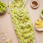A long platter filled with a creamy green dip, reminiscent of high protein pasta sauce, garnished with edamame and red pepper flakes. Surrounding it are bowls of edamame, spicy red pepper flakes, zesty lemon wedges, and crushed garlic on a beige background.