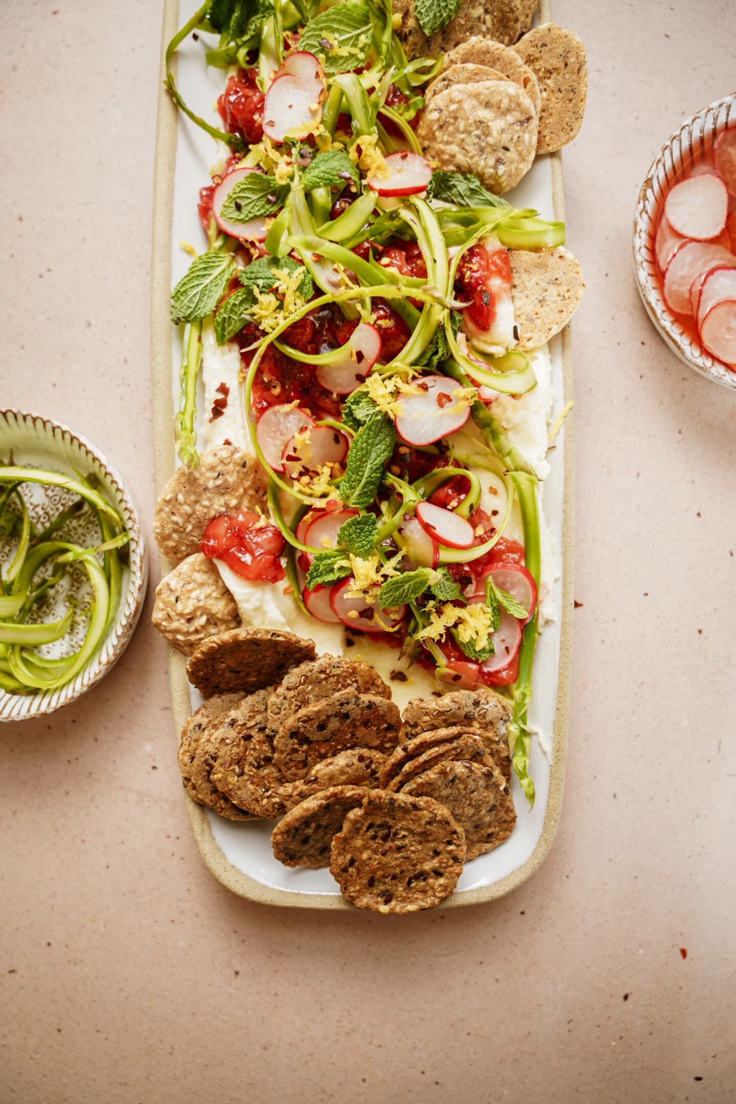 A rectangular platter of salad featuring sliced tomatoes, shaved asparagus, radishes, topped with herbs. Served with crisp crackers. Additional bowls with radish slices and shaved asparagus surround the platter on a light table.