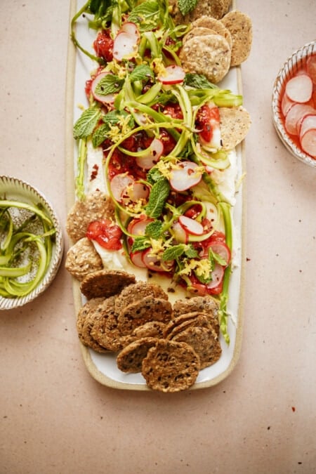 A rectangular platter of salad featuring sliced tomatoes, shaved asparagus, radishes, topped with herbs. Served with crisp crackers. Additional bowls with radish slices and shaved asparagus surround the platter on a light table.