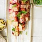 Skewers with grilled melon and pineapple chunks, garnished with fresh basil and lemon wedges, served on a white rectangular plate next to small bowls of lemon slices and basil leaves.