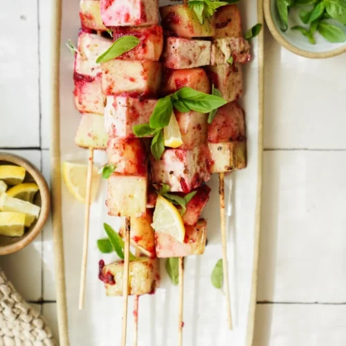 Skewers with grilled melon and pineapple chunks, garnished with fresh basil and lemon wedges, served on a white rectangular plate next to small bowls of lemon slices and basil leaves.