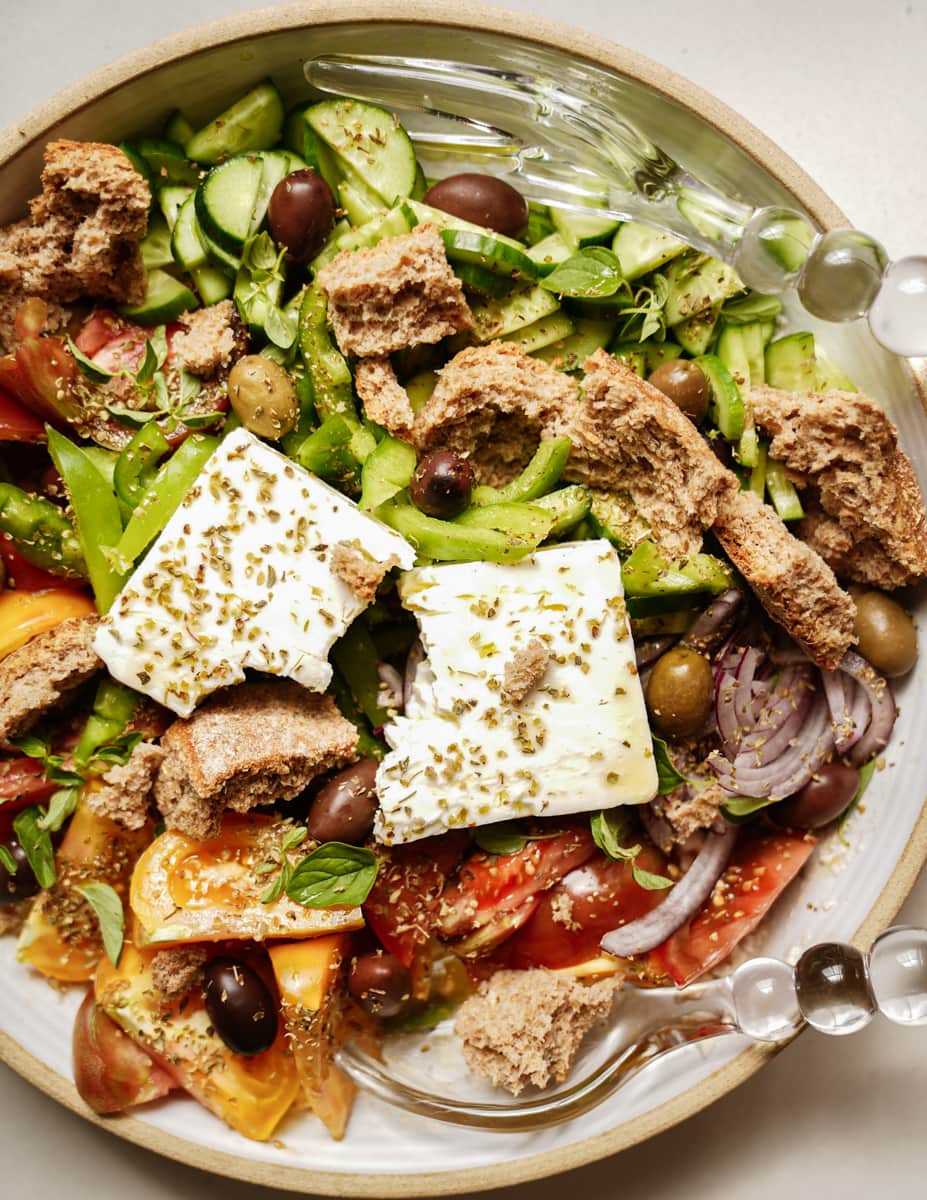 A colorful salad in a bowl with chopped cucumbers, tomatoes, red onions, black olives, herbs, crumbled bread, and two slices of feta cheese. Two clear serving utensils are placed in the bowl.