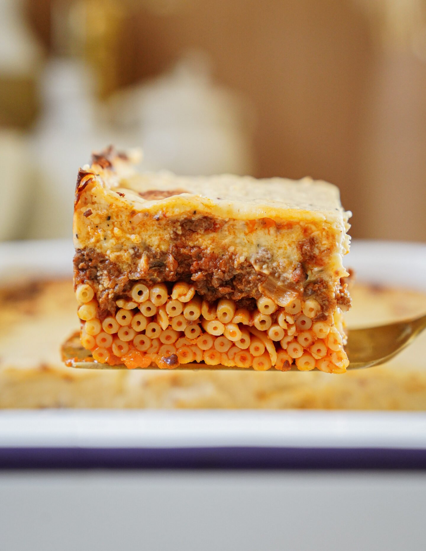 A close-up of a slice of baked pasta casserole with visible layers of tubular pasta, ground meat sauce, and creamy cheese, held on a spatula above a baking dish.