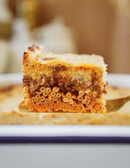 A close-up of a slice of baked pasta casserole with visible layers of tubular pasta, ground meat sauce, and creamy cheese, held on a spatula above a baking dish.