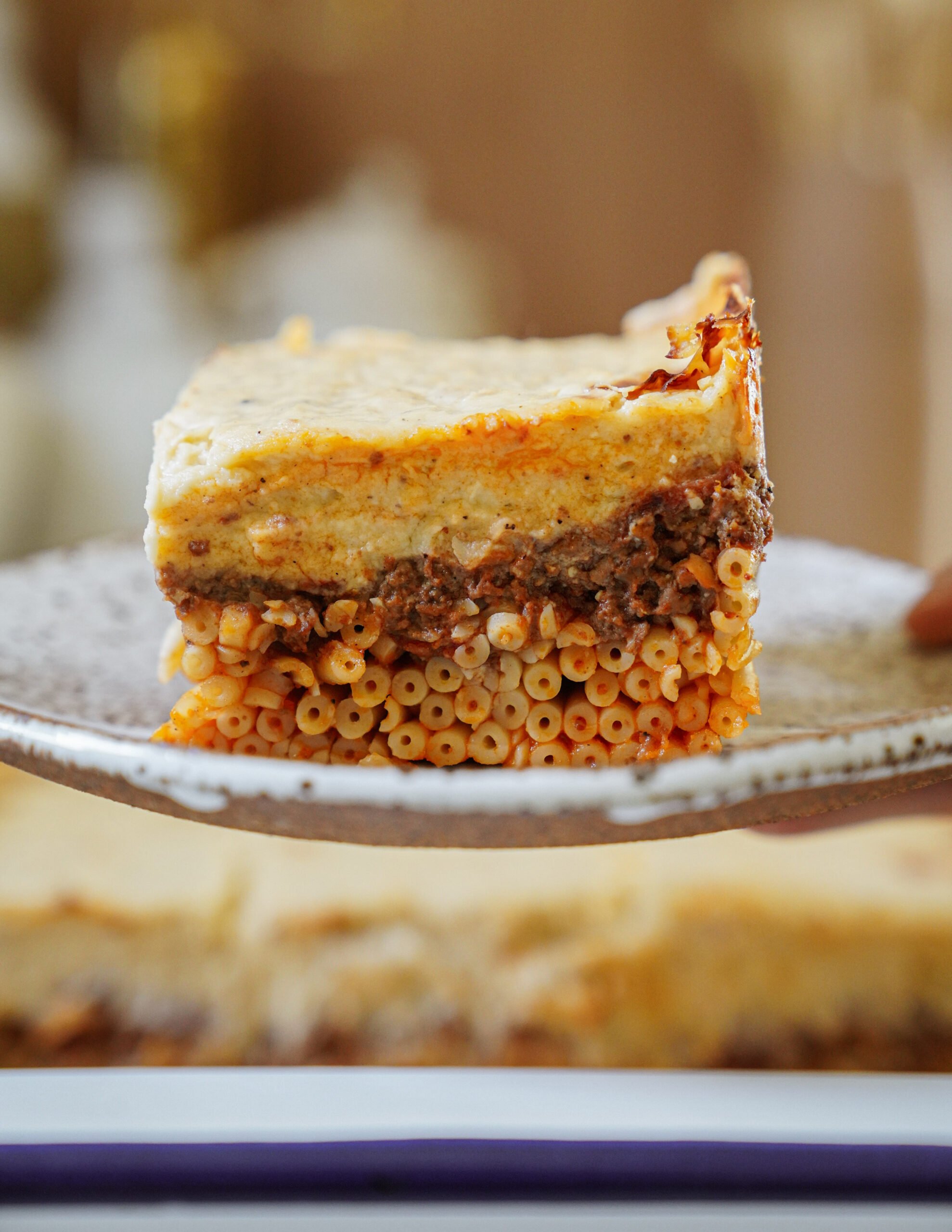 A close-up of a slice of pastitsio, a Greek casserole with layers of tubular pasta, seasoned ground meat, and creamy béchamel sauce, served on a ceramic plate.