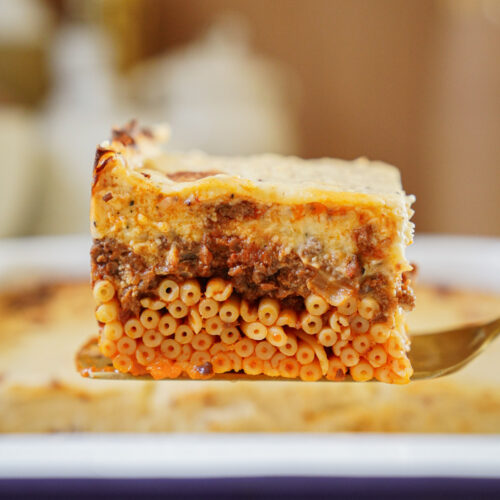 A close-up of a slice of baked pasta casserole with layers of small pasta tubes, ground meat sauce, and creamy cheese topping, held up on a spatula above a baking dish.