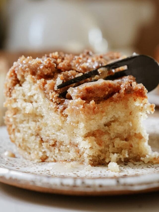 A slice of golden-brown coffee cake on a white plate, with a silver fork resting beside it. The cake's moist crumb is visible, topped with a generous layer of cinnamon-sugar streusel.