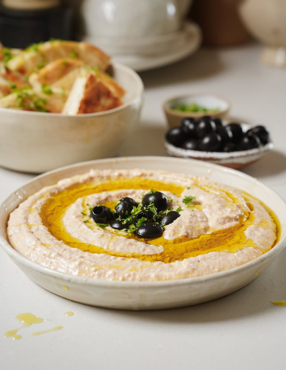 A bowl of hummus drizzled with olive oil and garnished with black olives and herbs sits on a table. In the background, theres a dish of sliced bread and a small bowl of black olives. The setting is a bright, inviting kitchen.