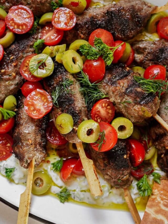Grilled meat skewers topped with cherry tomatoes, green olives, and fresh herbs are served on a plate. A creamy sauce is visible underneath, with bread on the side.