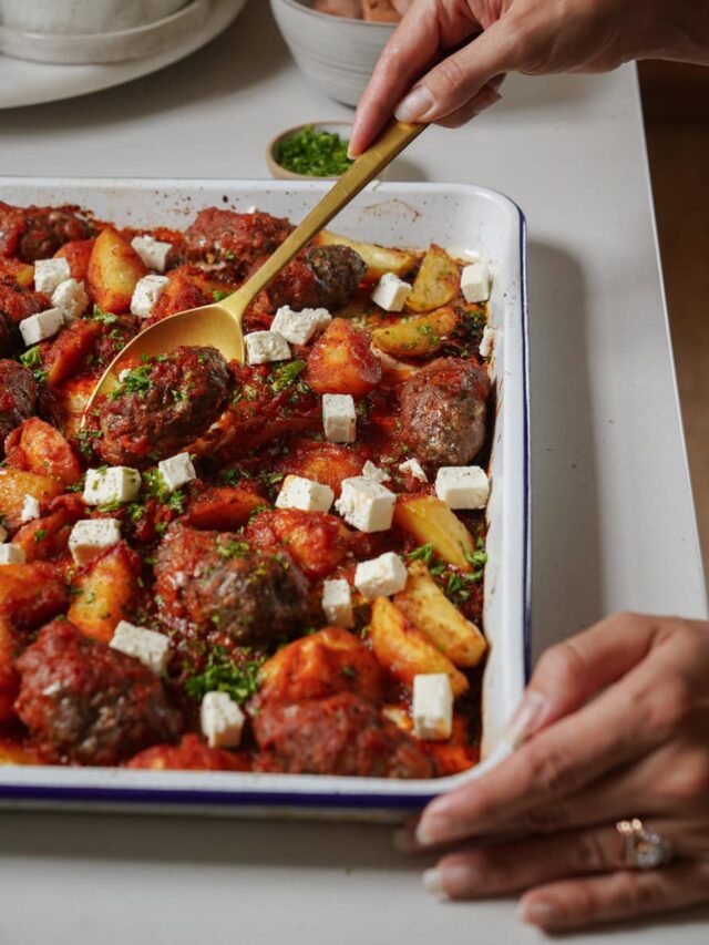 A person serves a baked dish with seasoned meatballs, potato wedges, tomato sauce, and cubes of feta cheese from a rectangular dish. A sprinkle of herbs garnishes the meal, and a small bowl of chopped herbs is visible in the background.