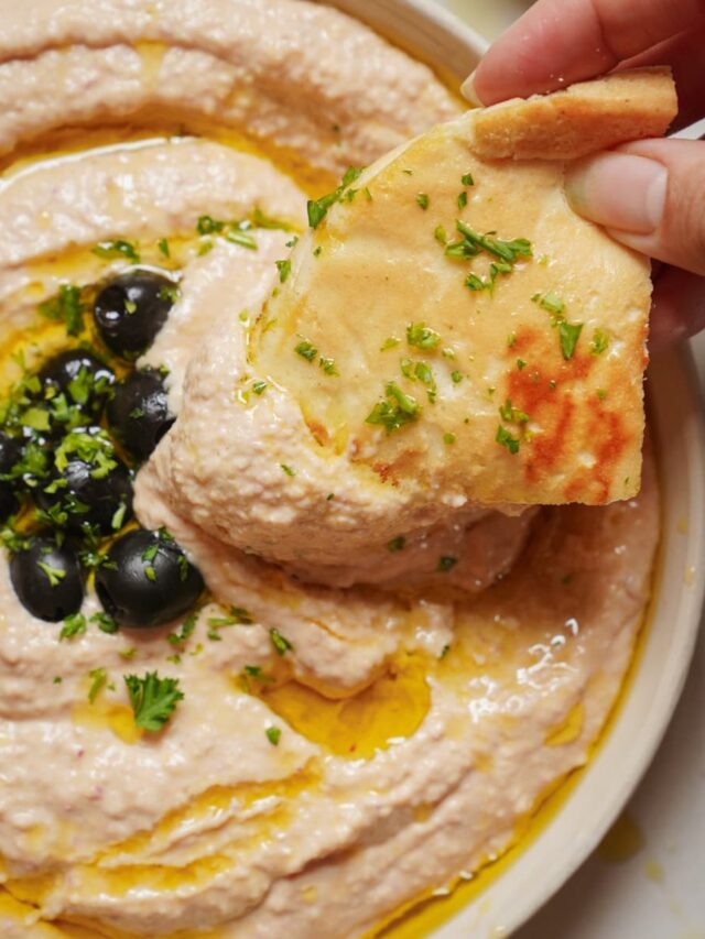 A close-up of a bowl of creamy dip garnished with black olives and chopped herbs. A hand is dipping a piece of flatbread, sprinkled with herbs, into the dip. Drizzles of olive oil are visible on the surface.