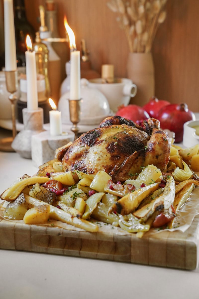 A roasted chicken on a wooden board surrounded by roasted potatoes and vegetables. Candles and ceramic vases are in the background, along with a pomegranate, creating a warm and rustic setting.